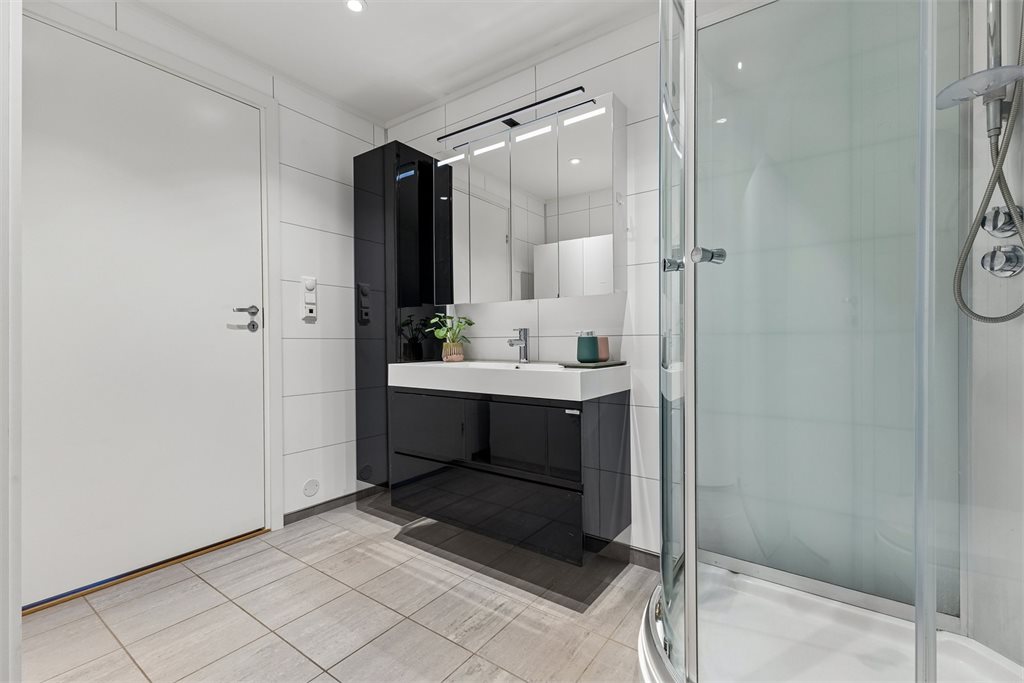 Badet fremstår moderne med flislagte overflater, vegghengt toalett, servantinnredning og dusjkabinett.