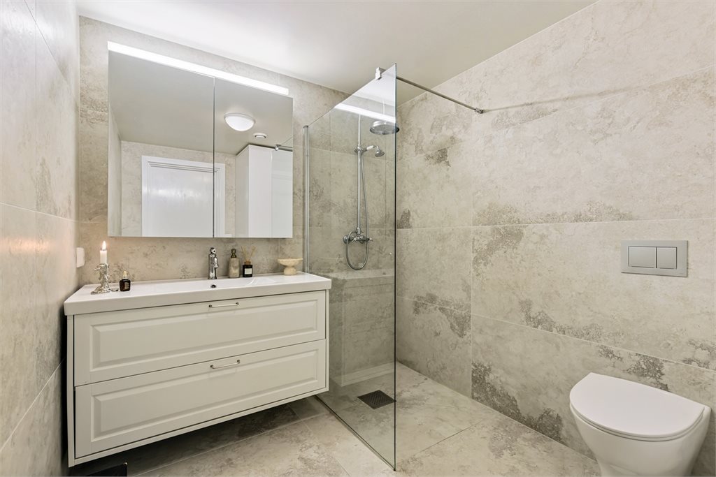 Badet er innredet med nedfelt servant, veggmontert toalett, dusjkabinett og opplegg for vaskemaskin. Gulvet er fliselagt og har varmekabler.